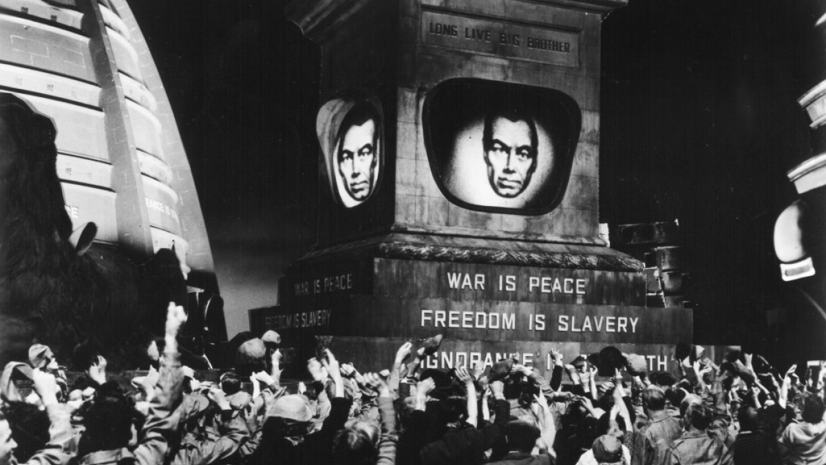 A black and white screenshot from an adaptation of 1984, showing Big Brother on the screen as crowds cheer in the foreground.