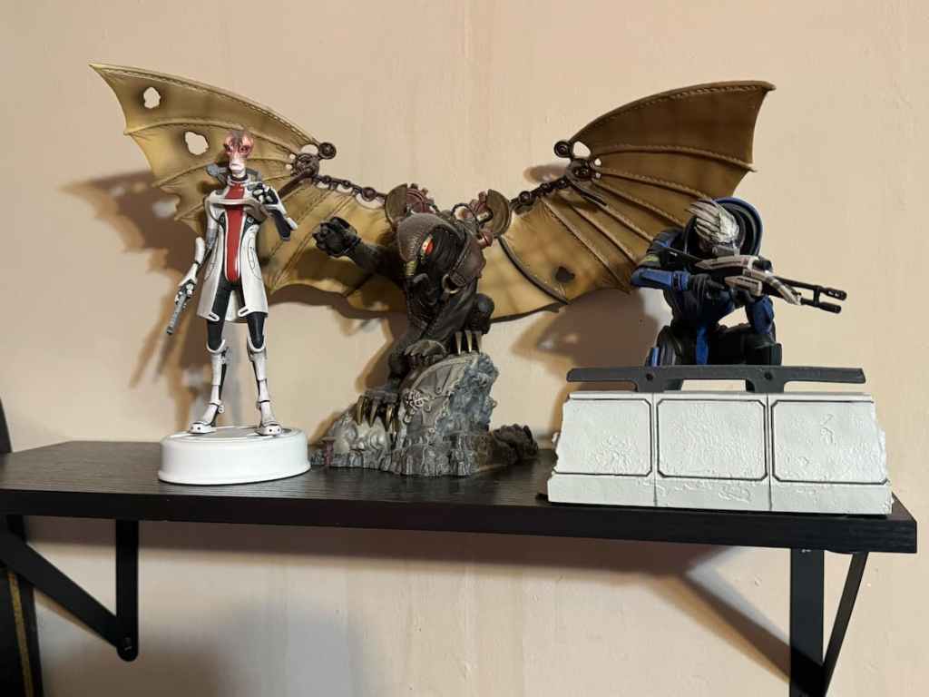 A shelf holding statues of (L-R) Mordin from Mass Effect, Songbird from BioShock Infinite, and Garrus from Mass Effect