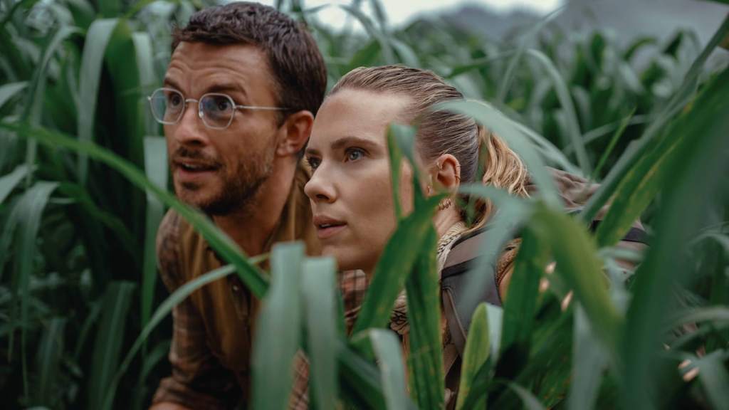A man wearing glasses with brown hair next to a blonde woman standing in tall grass in jurassic world rebirth