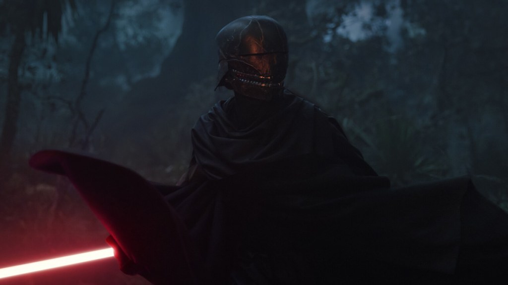 Qimir wearing a dark helmet with a creepy toothy smile and whirling around in battle holding a red lightsaber in The Acolyte